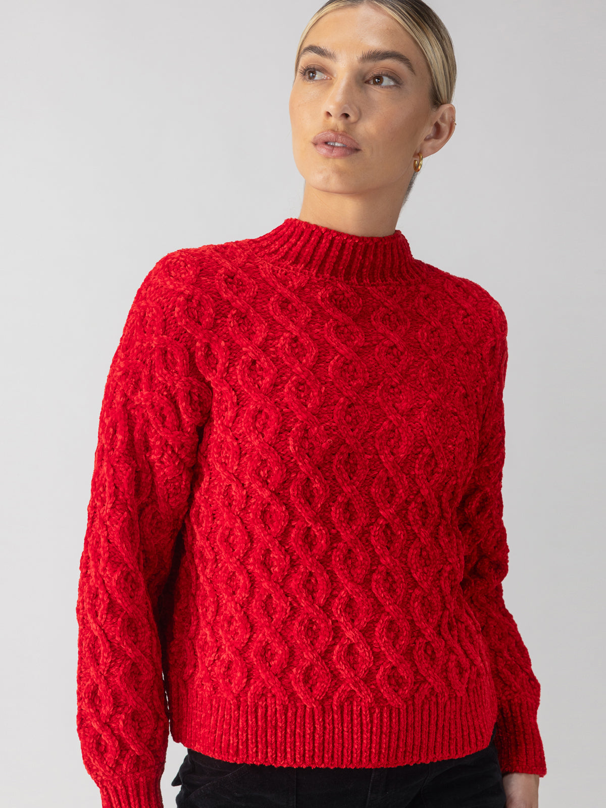 A person wearing the Cable Mock Neck Sweater in Mars Red by Sanctuary Clothing stands against a plain background. They have blond hair pulled back and are gazing off to the side. The sweater boasts a textured pattern along with ribbed cuffs and hem.