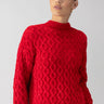 A person wearing the Cable Mock Neck Sweater in Mars Red by Sanctuary Clothing stands against a plain background. They have blond hair pulled back and are gazing off to the side. The sweater boasts a textured pattern along with ribbed cuffs and hem.