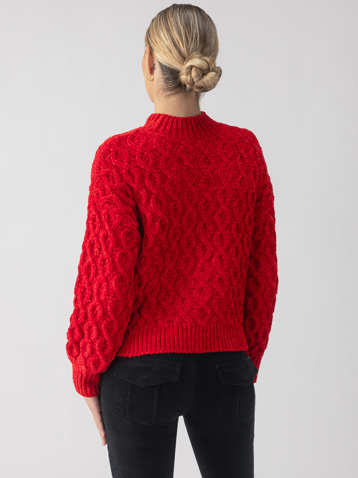 A person with blonde hair styled in a bun is wearing the Cable Mock Neck Sweater in Mars Red by Sanctuary Clothing, paired with black pants. They are facing away from the camera against a solid, light gray background.