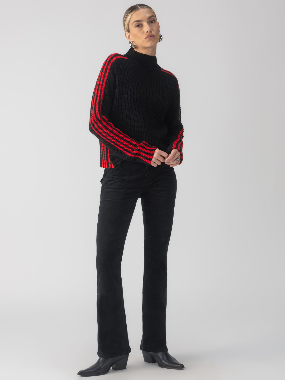 A person stands against a plain background, wearing the Sporty Stripe Mock Sweater in Black Multi from Sanctuary Clothing, black pants, and black boots. They have shoulder-length hair and are looking directly ahead.