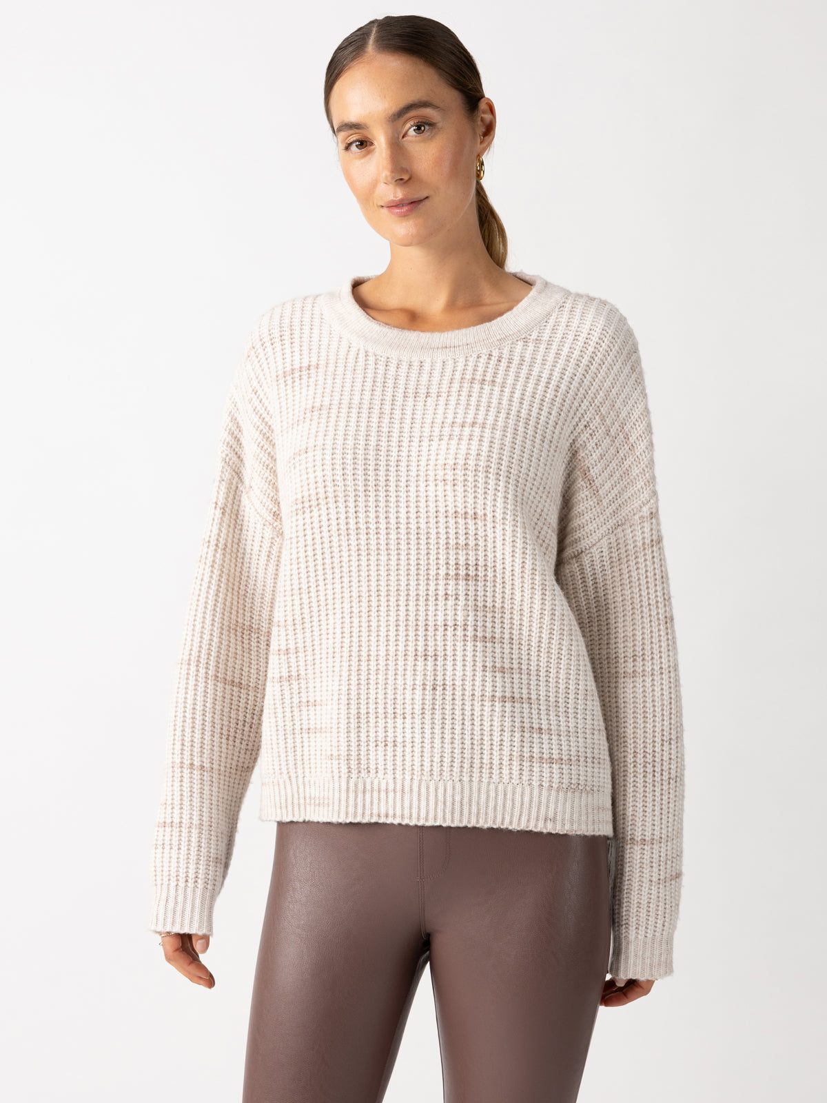 A person with long hair tied back is wearing a Sanctuary Clothing Cozy V Back Sweater in Coco Smoke Multi, paired with brown leather pants. The background is plain white.