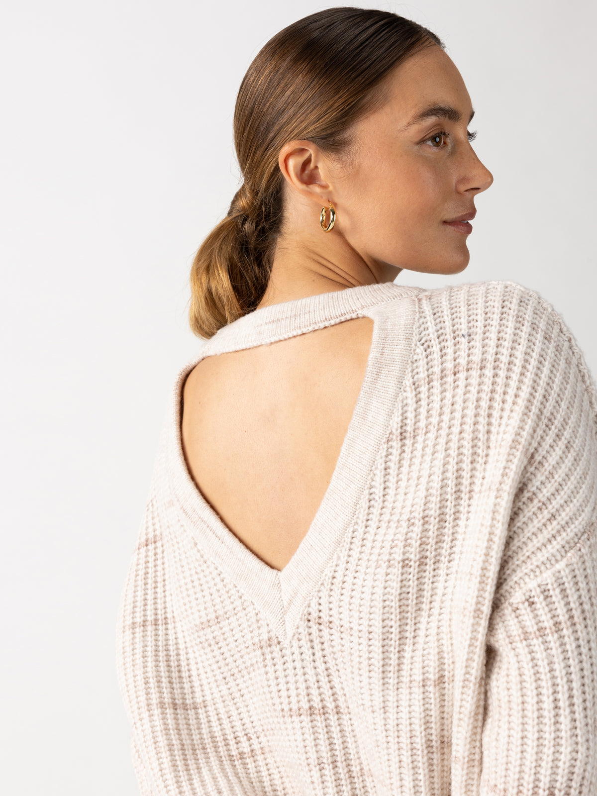 A person with hair pulled back into a low bun is wearing the Cozy V Back Sweater in Coco Smoke Multi by Sanctuary Clothing and gold hoop earrings, facing slightly to the side. The texture of the sweater is ribbed, and the background is neutral.