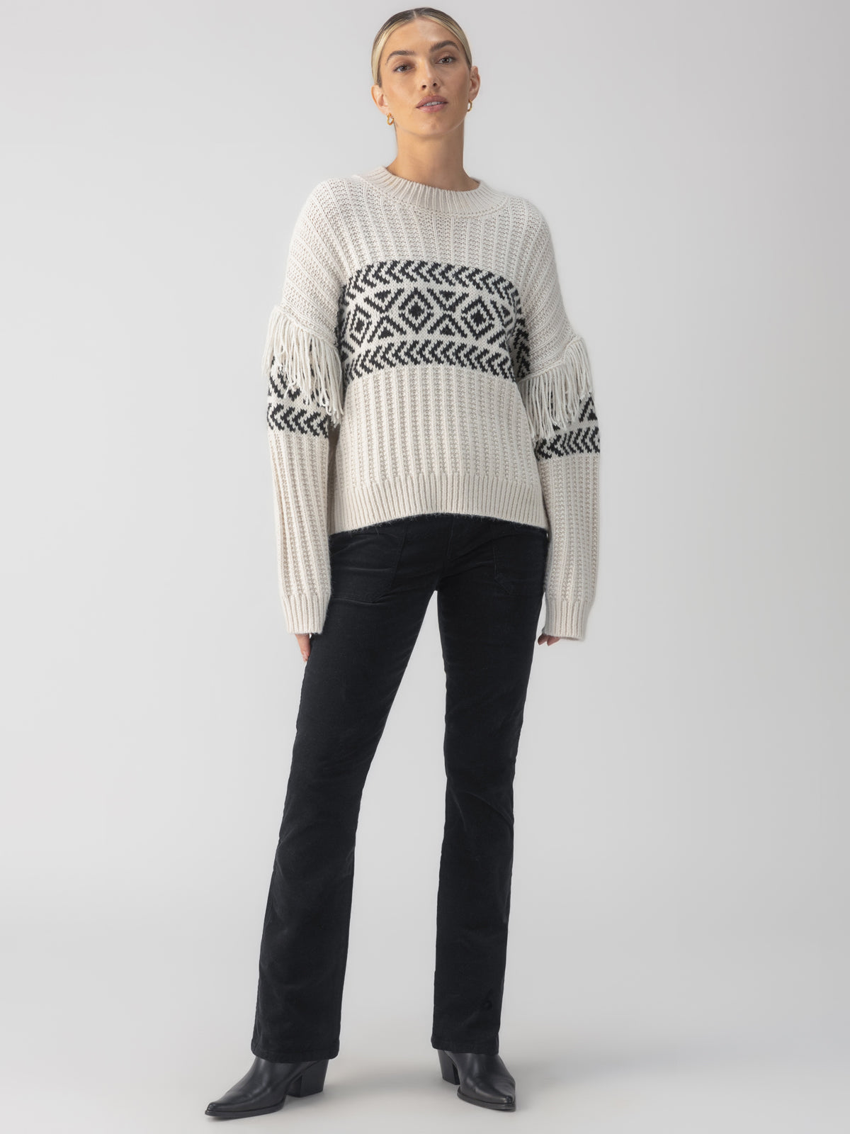 A person poses wearing the Artisan Fringe Sweater in Chalk and Black by Sanctuary Clothing, paired with black pants and black heeled shoes. They stand against a plain gray background.