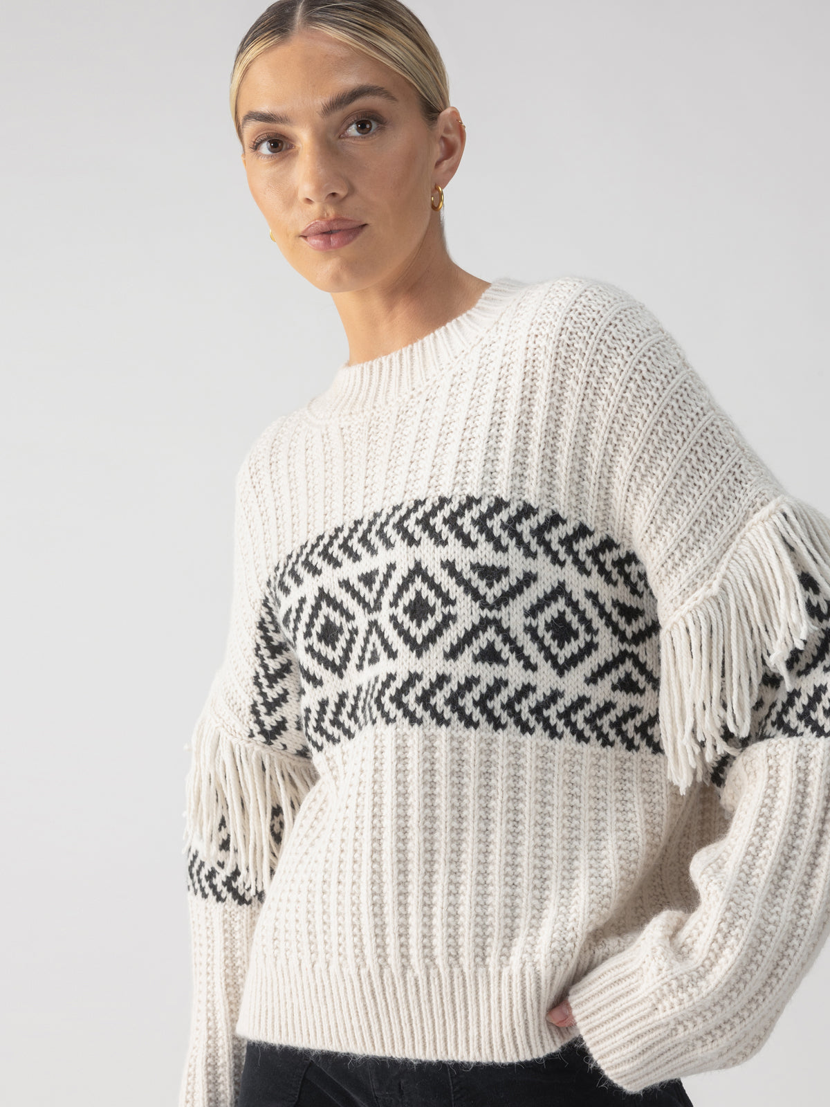 A person poses in the Artisan Fringe Sweater Chalk And Black by Sanctuary Clothing, set against a plain grey background. They have light hair pulled back and are wearing small hoop earrings.