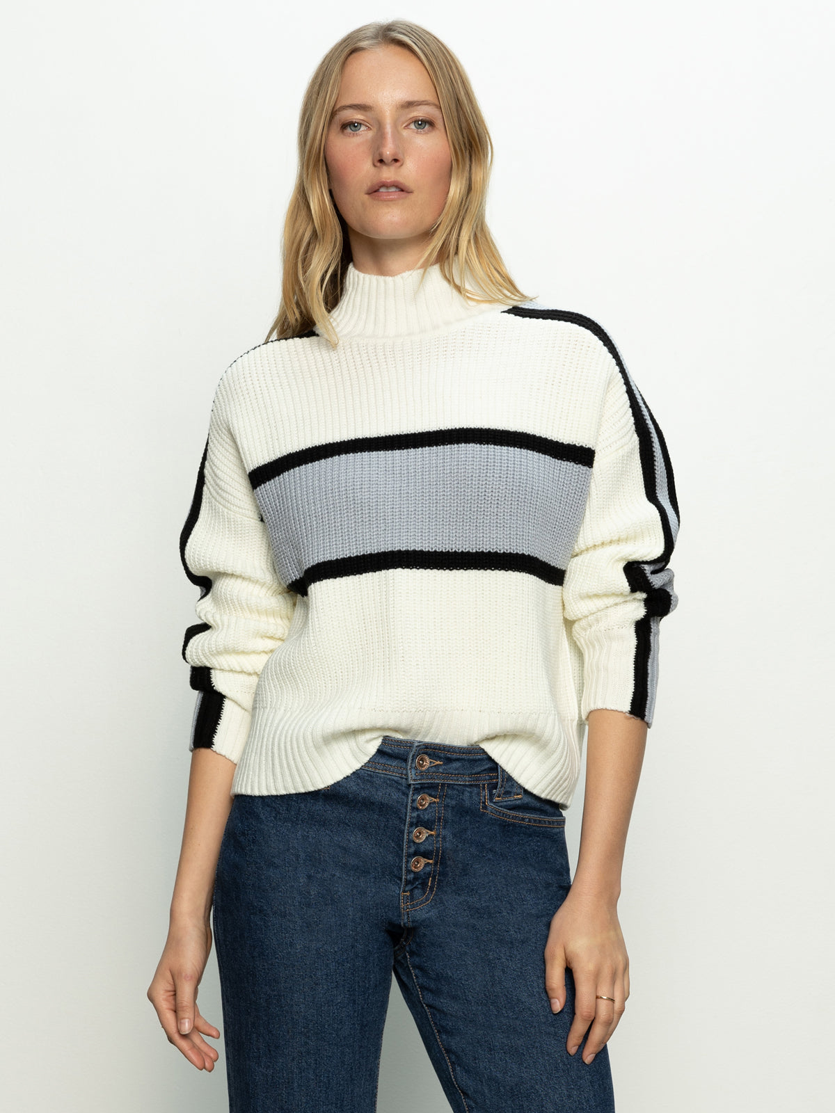 A person with long hair wears a Sanctuary Clothing's mock neck stripe sweater in pearl blue, black, and chalk stripes paired with blue jeans. They stand against a plain background with a confident expression and a casual pose.