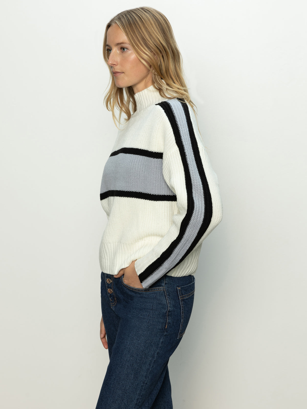 A person with long hair wears a Sanctuary Clothing mock neck stripe sweater in pearl blue, black, and chalk stripes. They're paired with blue jeans and stand against a plain light background.