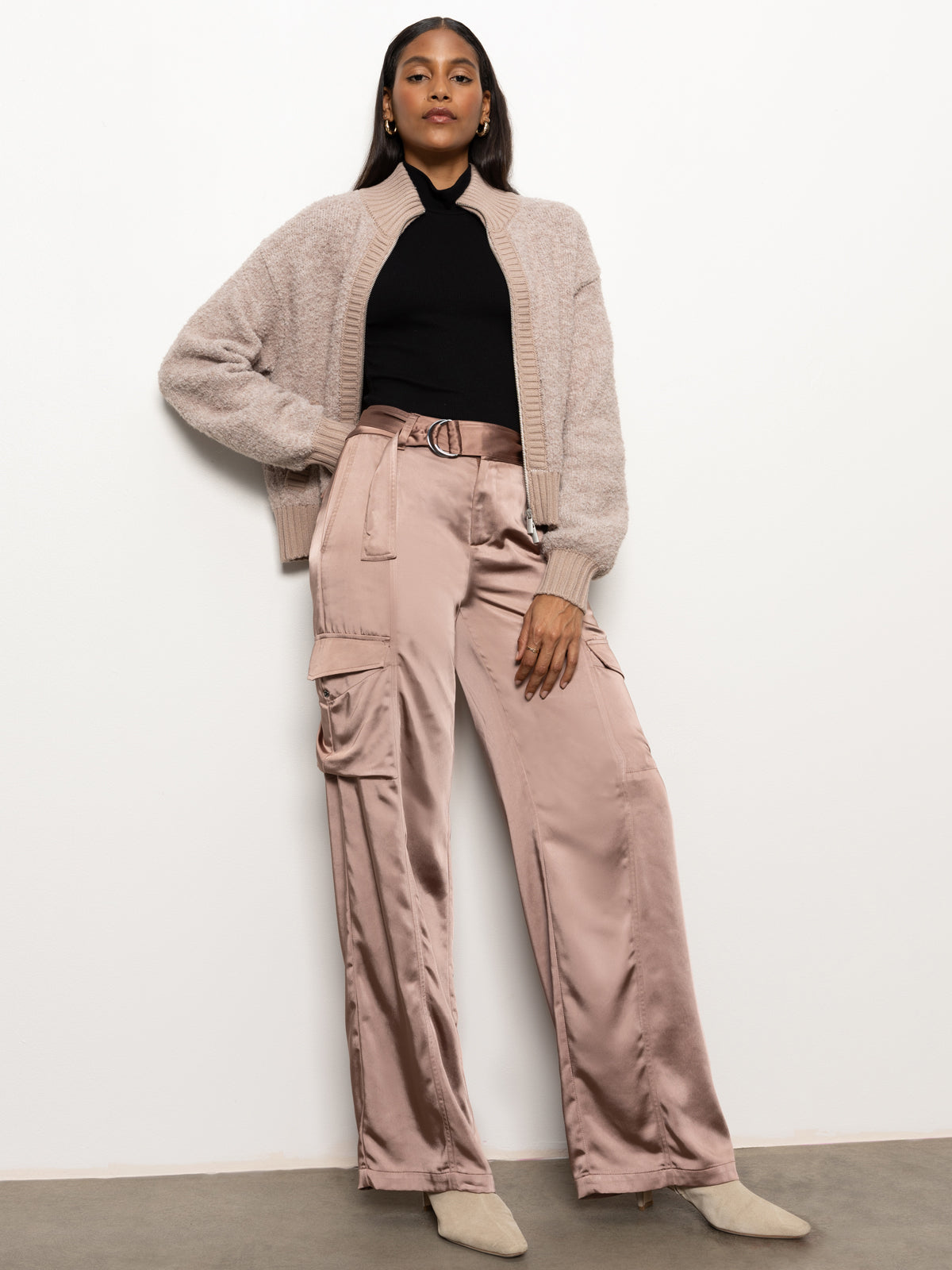 A person stands confidently against a white wall, wearing the "Boucle Knit Bomber Sweater Feather" by Sanctuary Clothing over a black top and satin pink cargo pants, with matching beige shoes. Their hands are in their pockets, and they are looking directly at the camera.