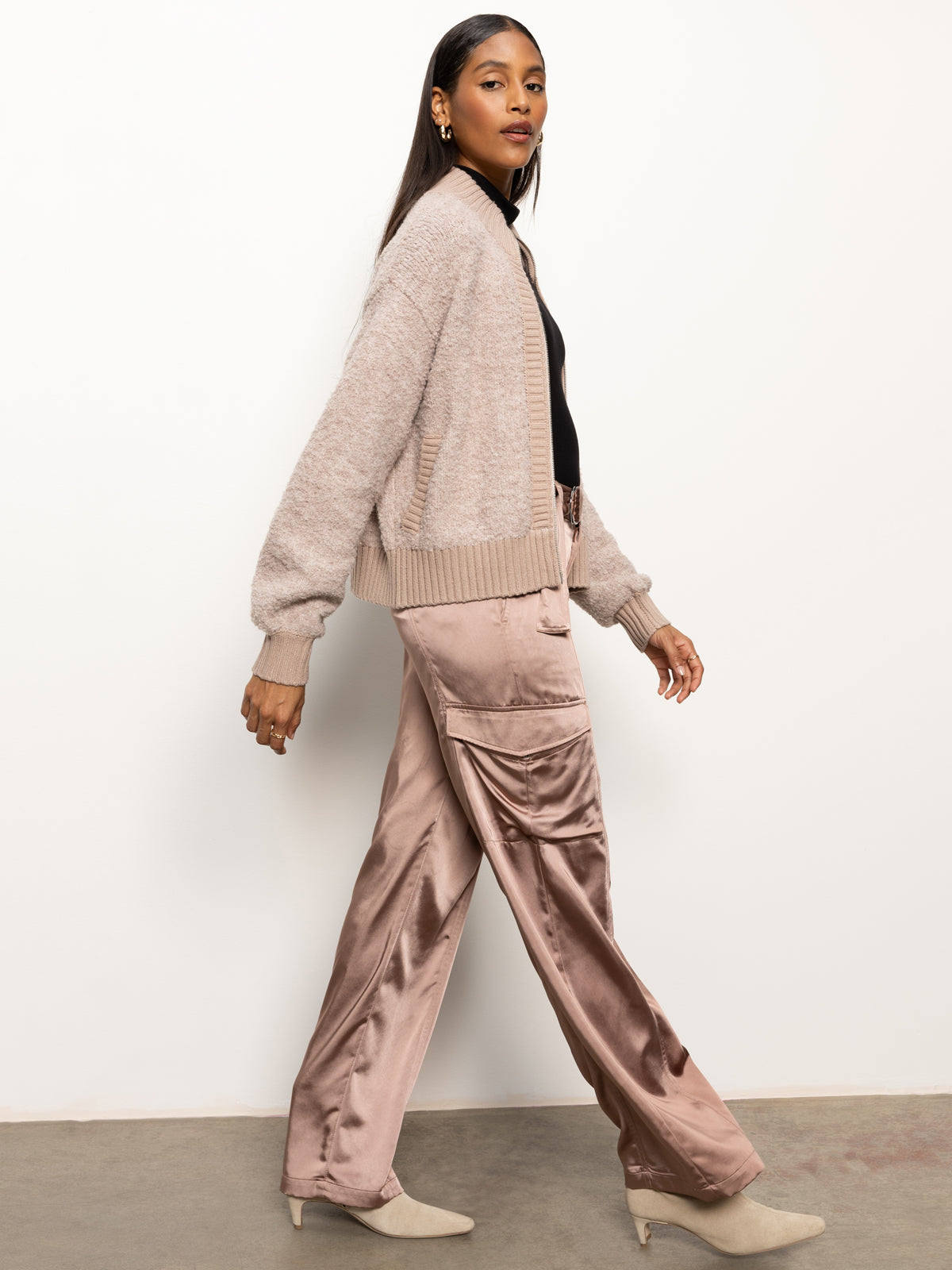 A woman in a boucle knit bomber sweater from Sanctuary Clothing and silky brown cargo pants strides confidently. She sports tan heeled boots and gold hoop earrings, her long hair cascading around her neutral expression, set against a plain white background.