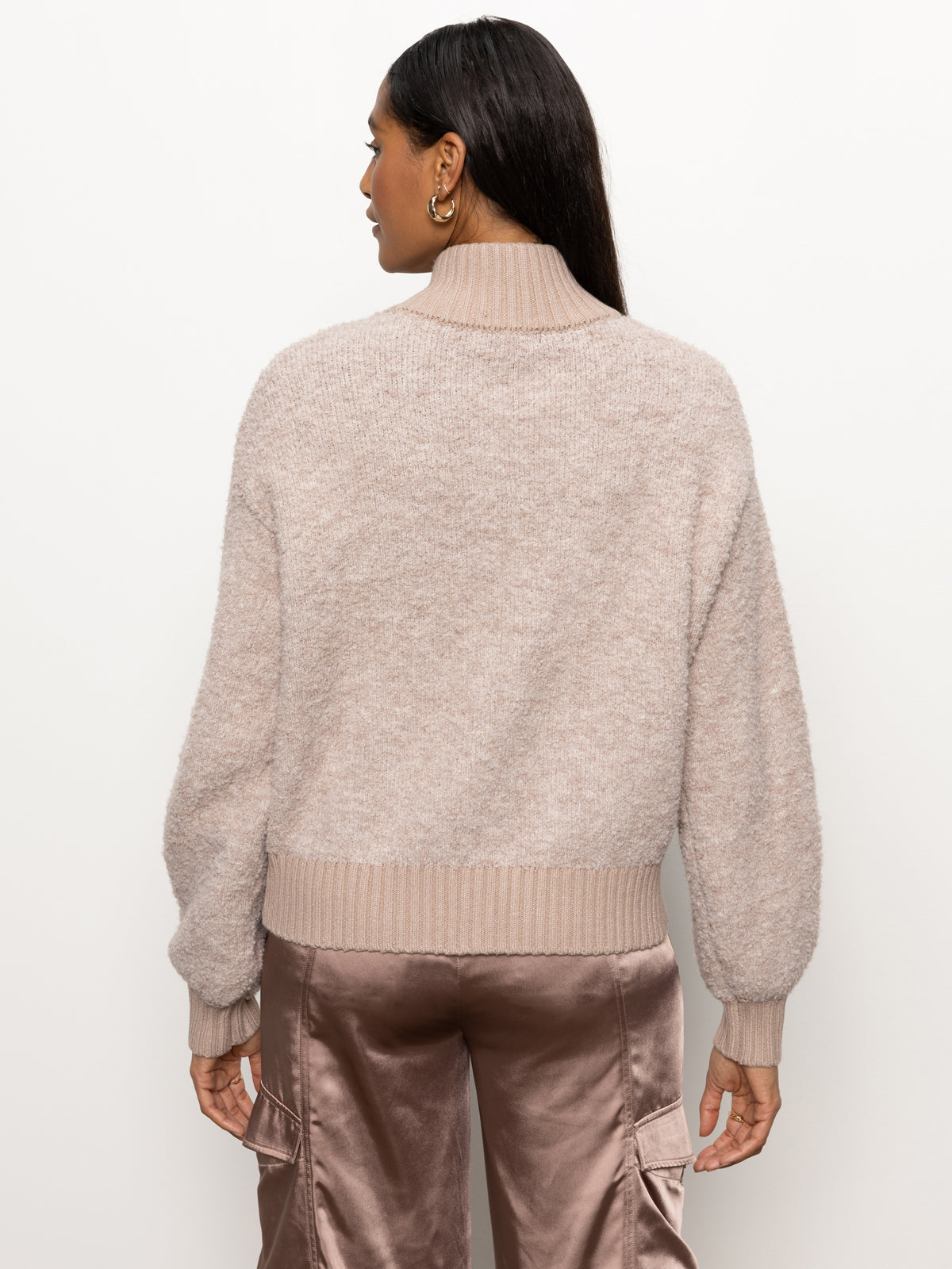 A woman with long dark hair is facing away, wearing a cozy boucle knit bomber sweater in beige by Sanctuary Clothing and satin cargo pants. She stands against a plain white background.