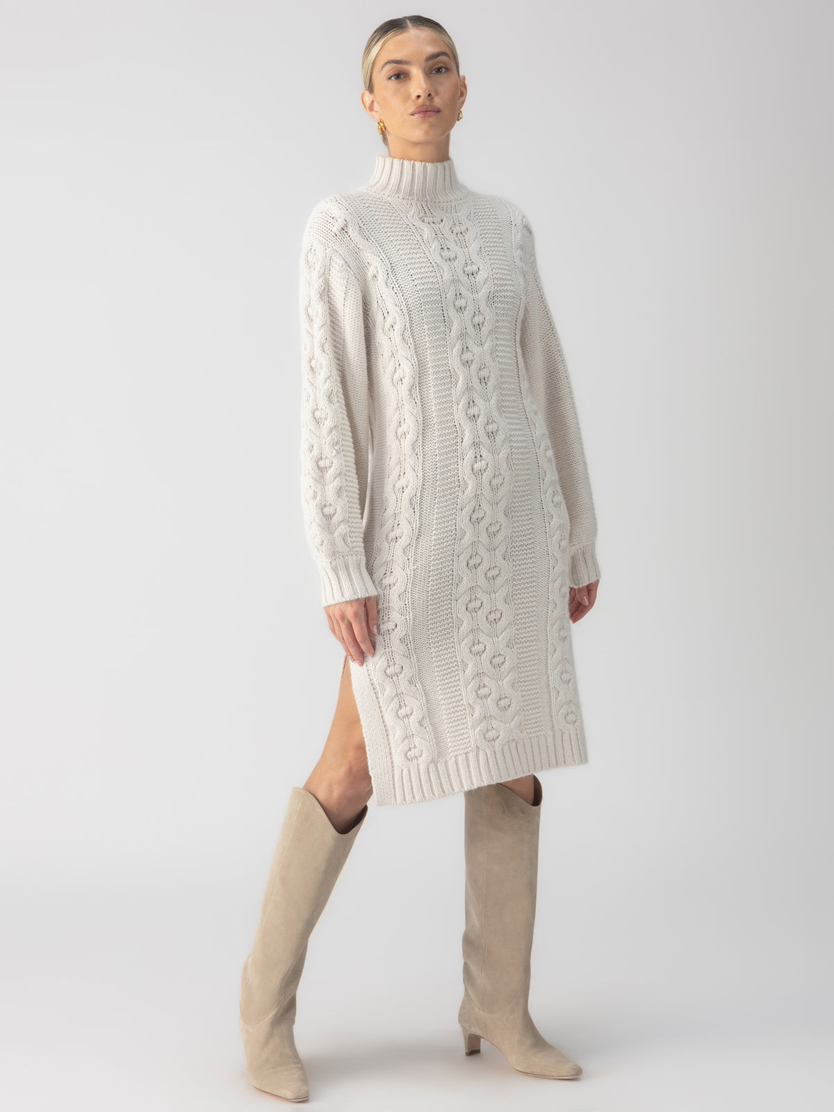 A person wearing the Cable Sweater Dress Chalk by Sanctuary Clothing, paired with tall beige boots, poses confidently against a plain background.