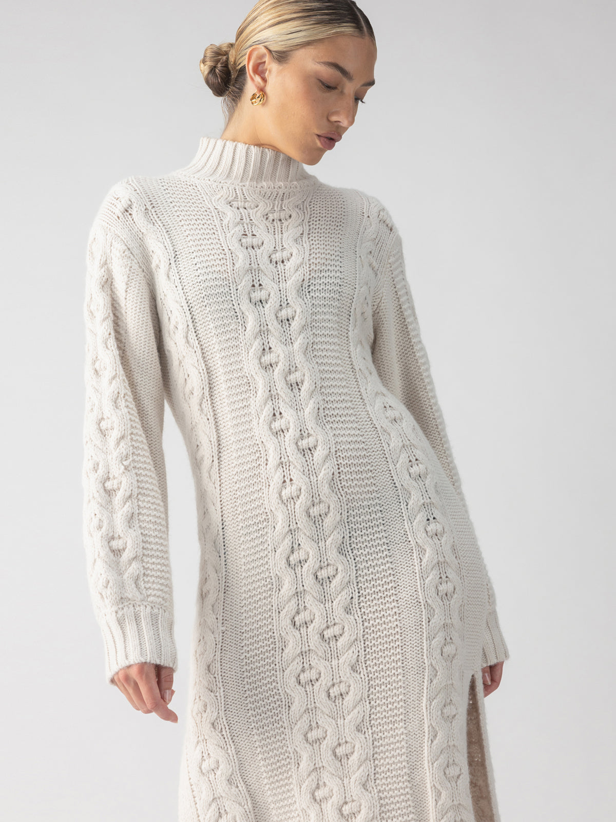 A person wearing the Cable Sweater Dress Chalk by Sanctuary Clothing stands against a light gray background. The cream-colored dress features cable-knit detailing, long sleeves, and a high collar, while the person's hair is styled in a low bun.