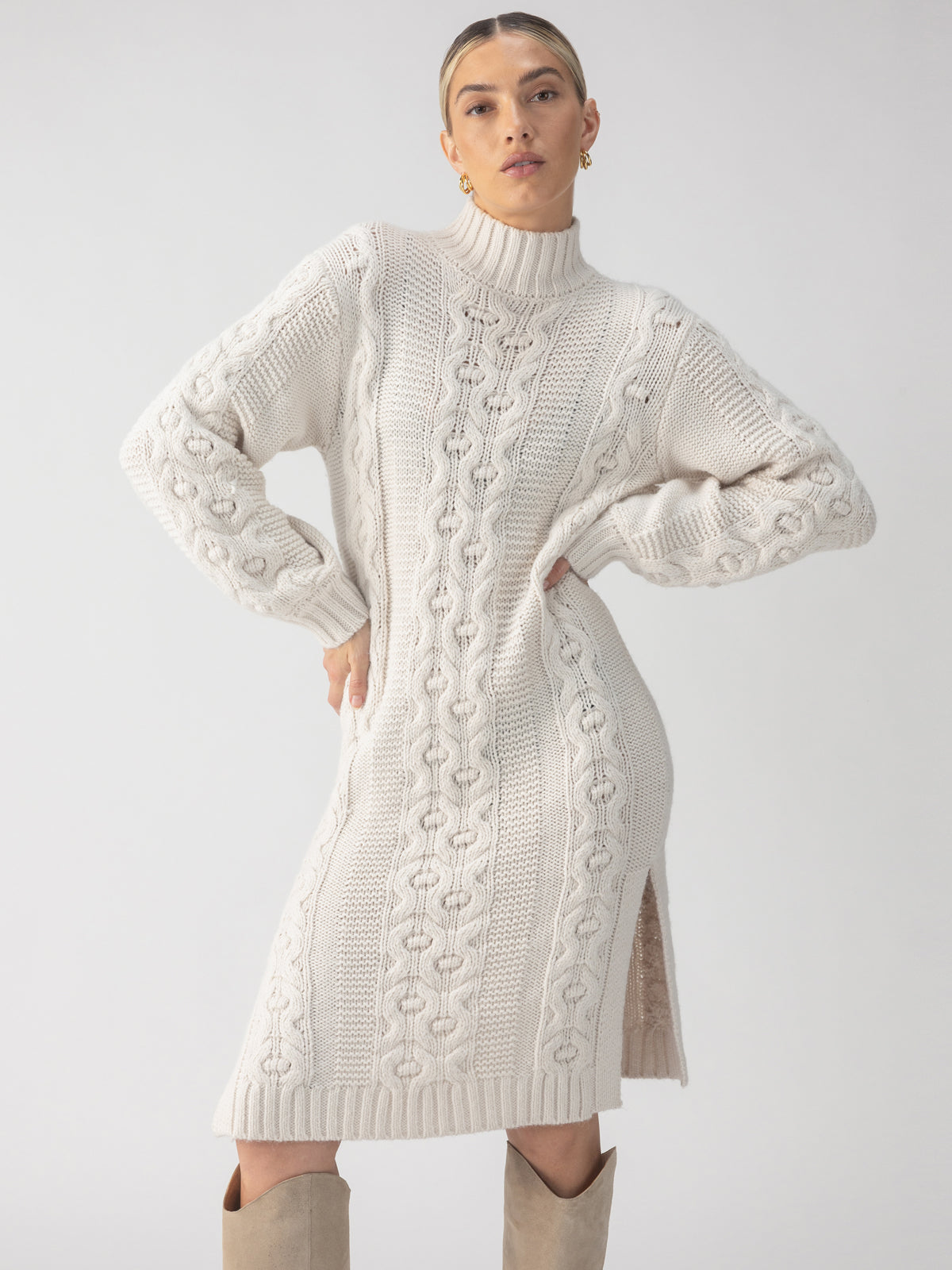 Dressed in the Cable Sweater Dress Chalk by Sanctuary Clothing, a person stands confidently with hands on hips. The cream-colored, cable-knit sweater dress features a high collar, long sleeves, and a side slit. They complete the look with knee-high beige boots.