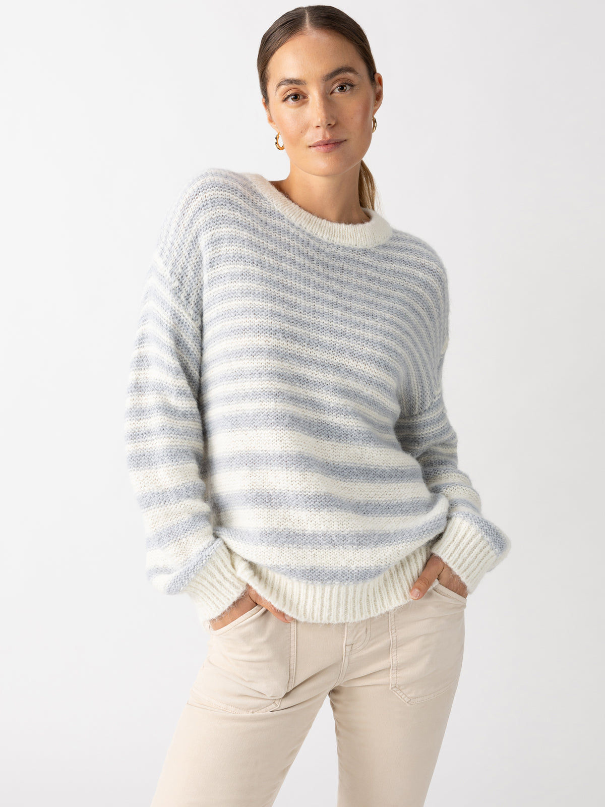 A person stands against a plain background, wearing Sanctuary Clothing's comfy, oversized Fuzzy Tunic Sweater in Pearl Blue Chalk Stripe. They have their hands in the pockets of light beige pants, giving off a relaxed and casual vibe.