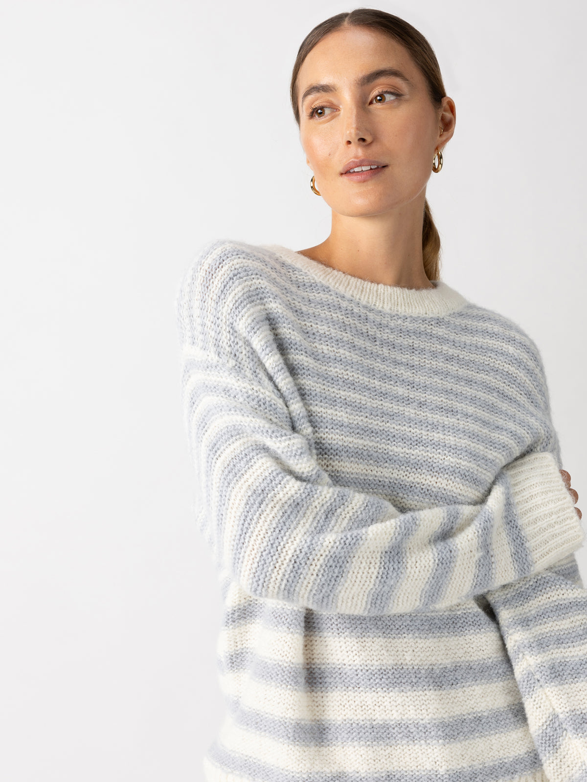 A person wearing the Sanctuary Clothing Fuzzy Tunic Sweater in Pearl Blue Chalk Stripe looks to the side against a plain white background. They have long hair pulled back and are adorned with small hoop earrings.