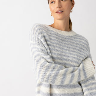 A person wearing the Sanctuary Clothing Fuzzy Tunic Sweater in Pearl Blue Chalk Stripe looks to the side against a plain white background. They have long hair pulled back and are adorned with small hoop earrings.