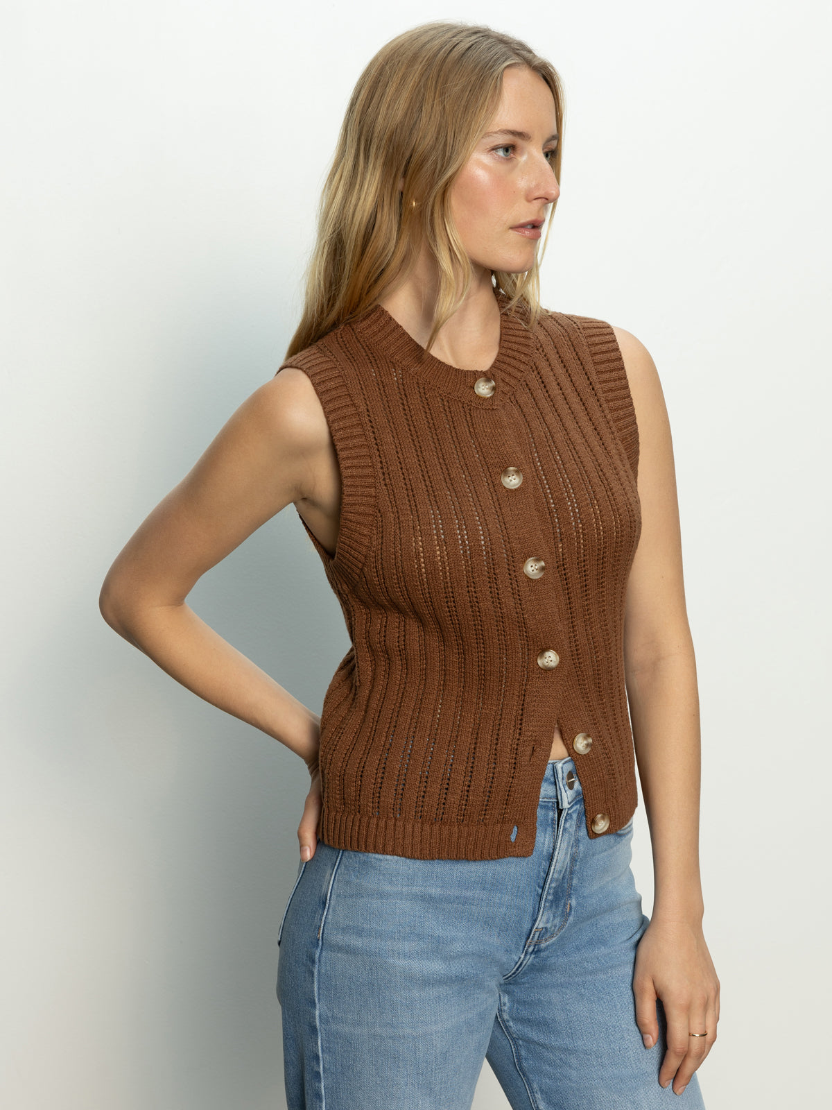 A woman with long blonde hair wears the Sanctuary Clothing pointelle shell sepia, a sleeveless button-up brown knit top, paired with light blue jeans. She stands against a plain white background, looking left with her right hand on her hip.