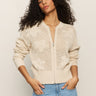 A person with long, curly hair wears a flower pop open stitch jacket in eco natural by Sanctuary Clothing, paired with light blue jeans. Their hands rest in their jean pockets as they stand against a plain background.