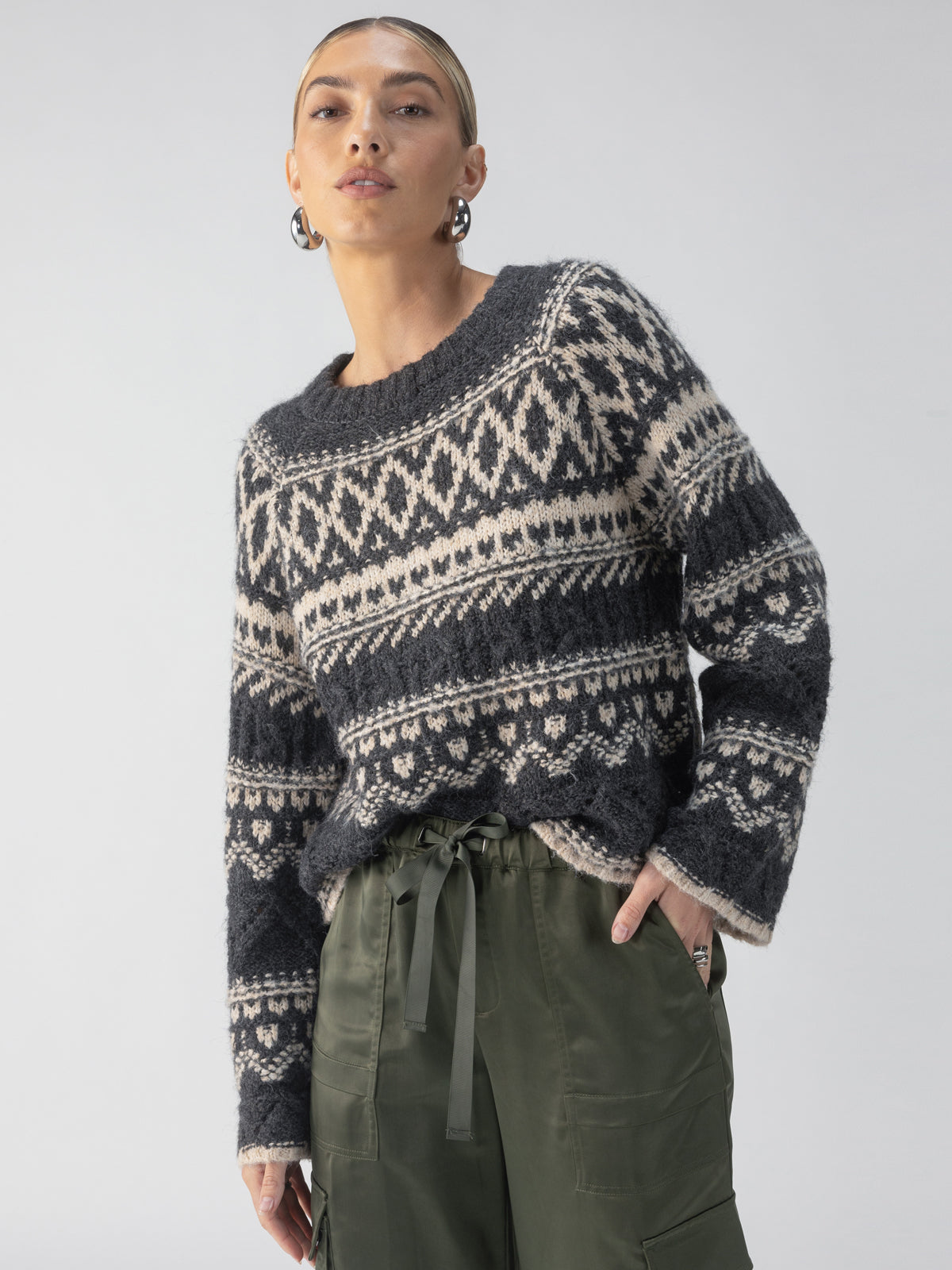 A person wearing the Sanctuary Clothing Fairisle Crew Neck Sweater in Heather Ash Multi stands with one hand in the pocket of their olive green cargo pants. They have short hair and are accessorized with hoop earrings, set against a plain, light-colored background.