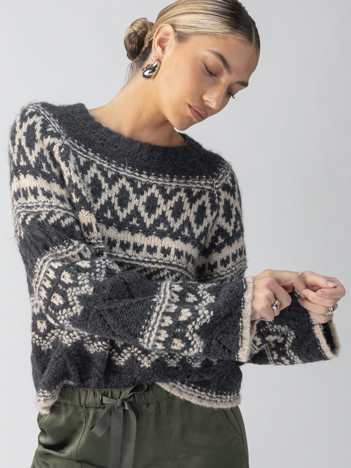 A woman with her hair in a low bun is wearing the Fairisle Crew Neck Sweater in Heather Ash Multi by Sanctuary Clothing. She accessorizes with large hoop earrings and adjusts her sleeve while pairing the sweater with green pants, all set against a plain background.