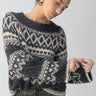 A woman with her hair in a low bun is wearing the Fairisle Crew Neck Sweater in Heather Ash Multi by Sanctuary Clothing. She accessorizes with large hoop earrings and adjusts her sleeve while pairing the sweater with green pants, all set against a plain background.