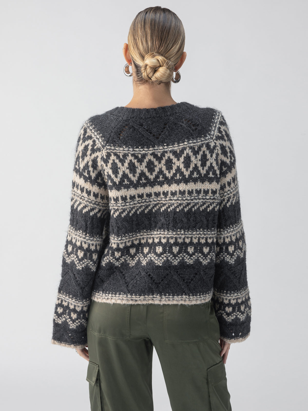 Rear view of a person with blonde hair in a bun, wearing the Fairisle Crew Neck Sweater Heather Ash Multi by Sanctuary Clothing, paired with green cargo pants. The sweater showcases intricate geometric designs as the person stands against a plain background.