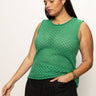 A woman in a Sanctuary Clothing open stitch shell sweater palm inclusive collection, paired with black pants, poses against a plain background. With long brown hair, she accessorizes with hoop earrings and layered necklaces, gazing downwards.