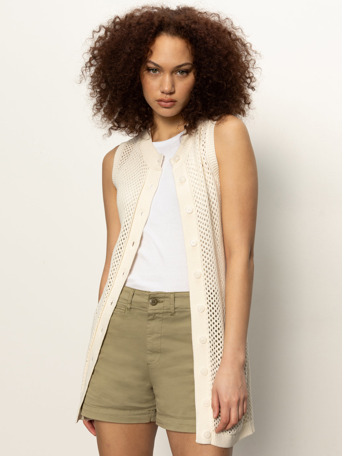 A woman with curly hair wears the Sanctuary Clothing long openwork vest macrame over a white top and olive green shorts, standing against a plain light background.