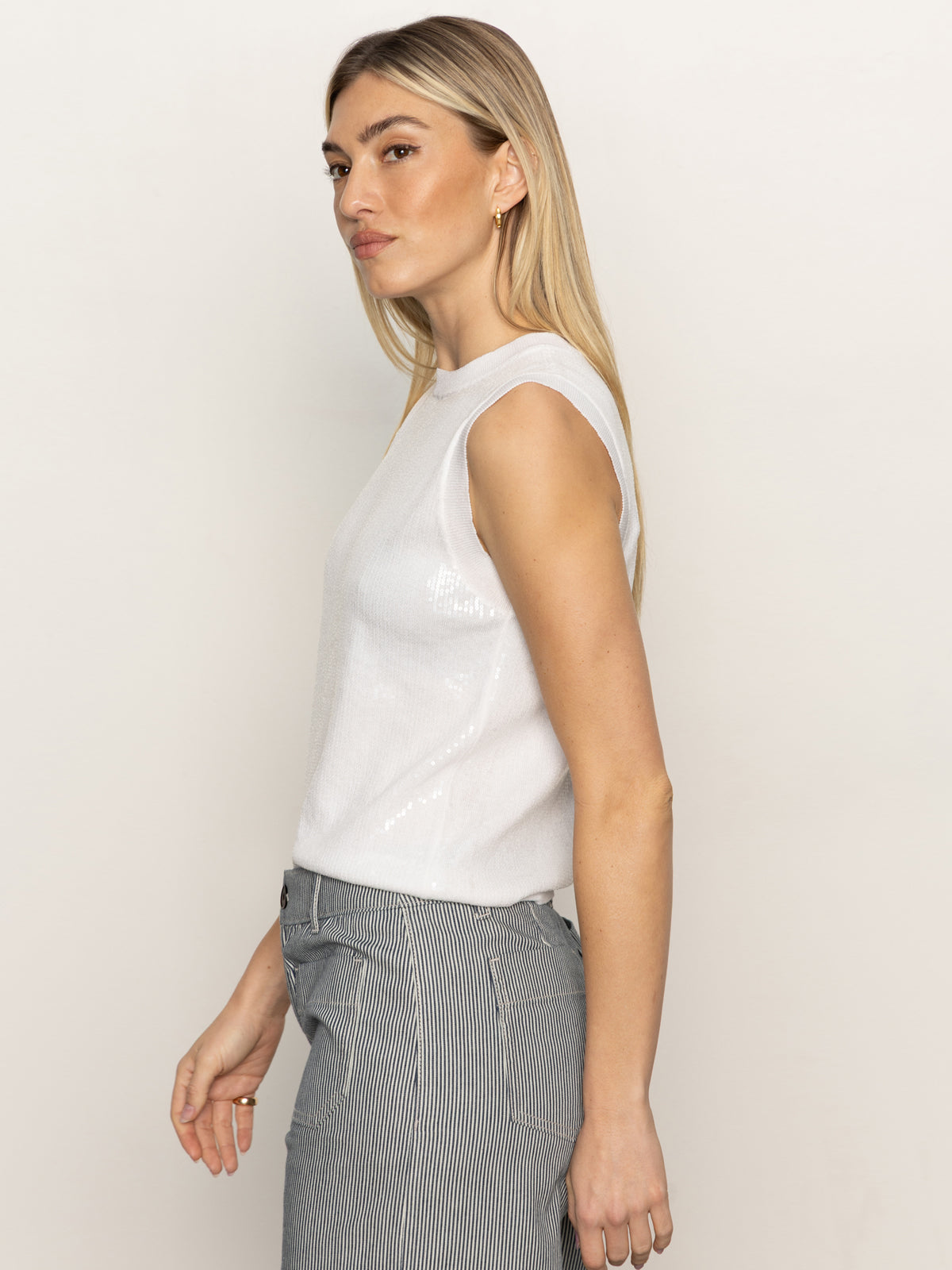 A woman with long blonde hair is wearing a Sanctuary Clothing sequin knitwear shell sweater in white, paired with striped gray pants, and standing in profile against a plain background.