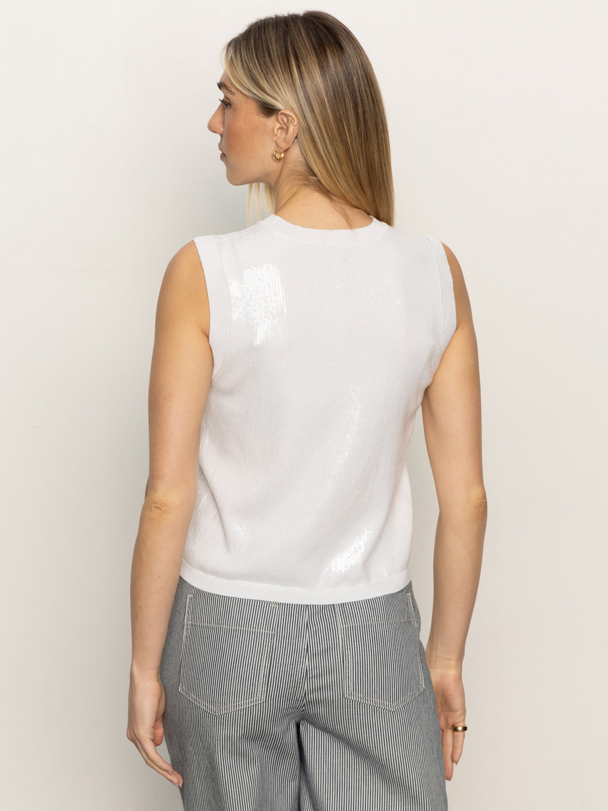 A woman with long blonde hair, wearing a white sequin knitwear shell sweater by Sanctuary Clothing and pinstriped gray pants, faces away from the camera against a plain light background.