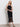 A woman with wavy blonde hair poses against a white wall wearing the Sanctuary Clothing promenade halter dress in black, featuring a fitted bodice, crochet details, fringe hem, and paired with black strappy sandals.