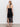 A woman with wavy blonde hair wears the Sanctuary Clothing promenade halter dress in black, a crochet backless style with a fringed hem and side slit, paired with black flat sandals. She stands facing away, highlighting the dress’s open back and details.