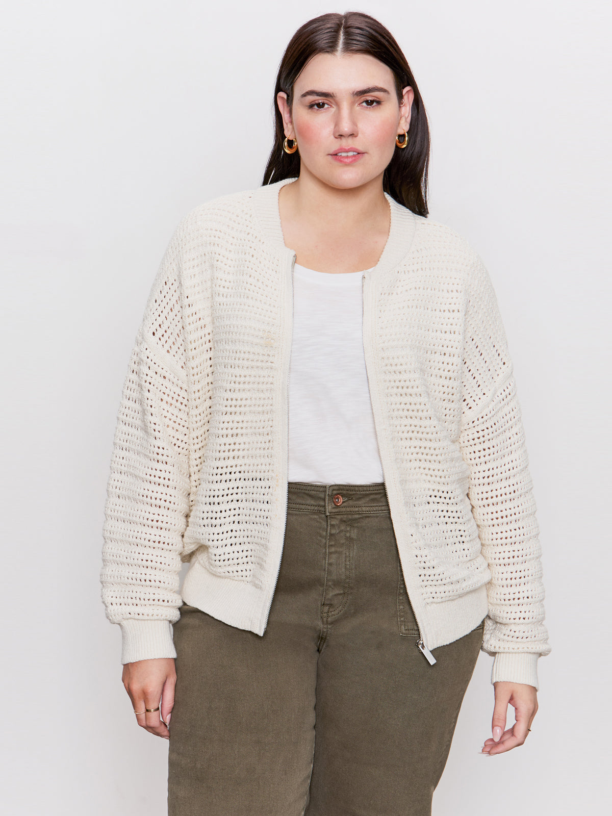 A person with long dark hair wears a cream-colored open knit bomber jacket from Sanctuary Clothing over a white shirt with olive green pants, standing against a plain white background and looking directly at the camera.