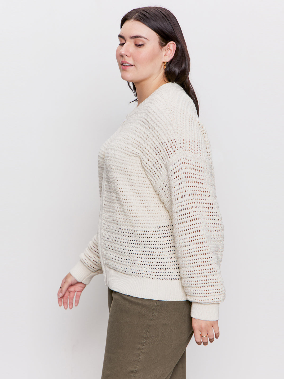A person with long, dark hair is standing side-on against a plain white background. They appear relaxed, wearing Sanctuary Clothing's open knit bomber jacket in chalk and olive green pants, looking slightly downward.
