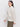 A person with long, dark hair is standing side-on against a plain white background. They appear relaxed, wearing Sanctuary Clothing's open knit bomber jacket in chalk and olive green pants, looking slightly downward.