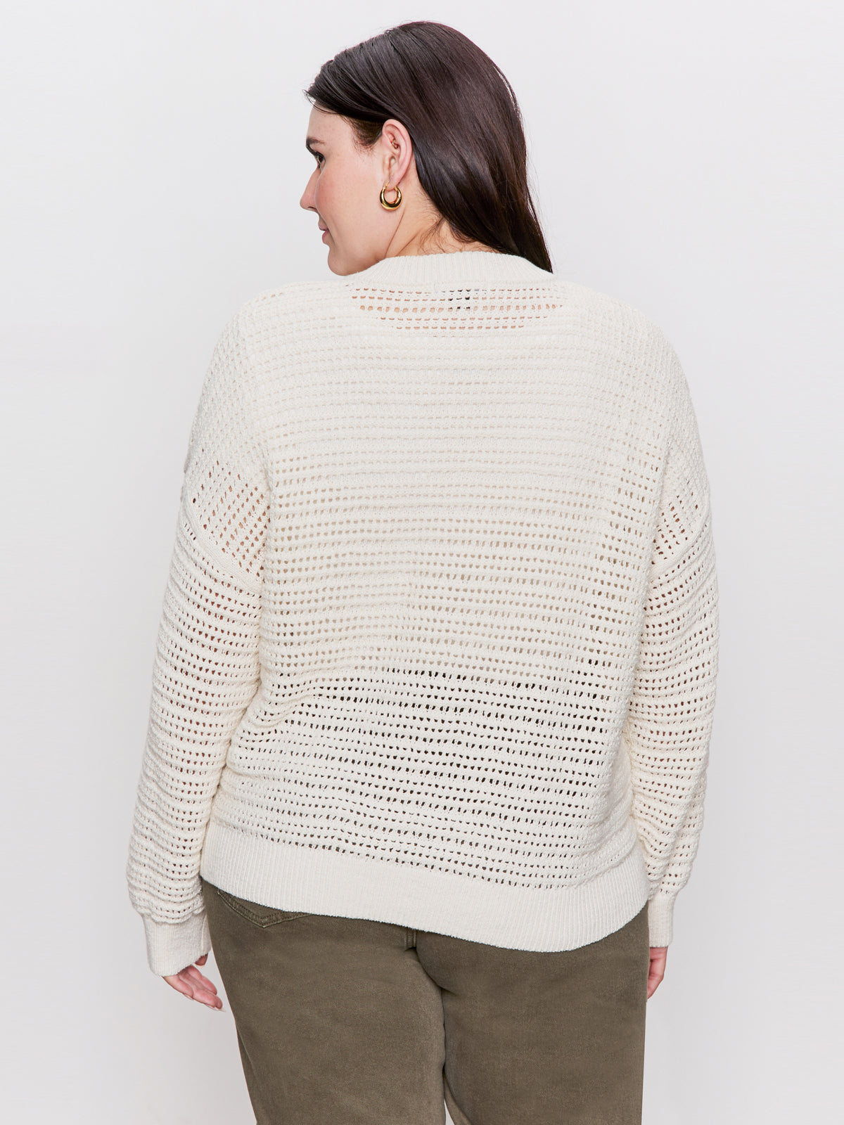 A woman, with dark brown hair and hoop earrings, faces away wearing a Sanctuary Clothing open knit bomber jacket in chalk. The background is plain white.