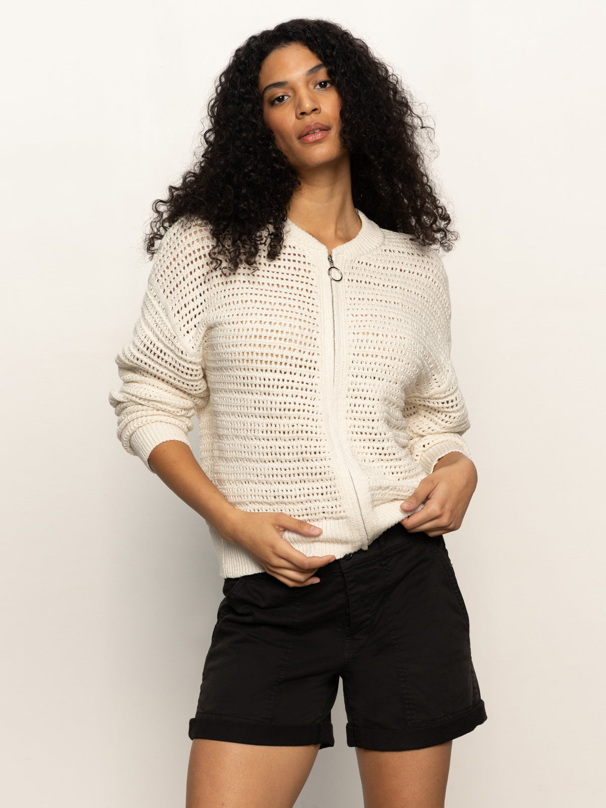 A person with long curly hair wears a Sanctuary Clothing open knit bomber jacket in chalk and black shorts, against a plain background. They stand relaxed, hands in pockets.