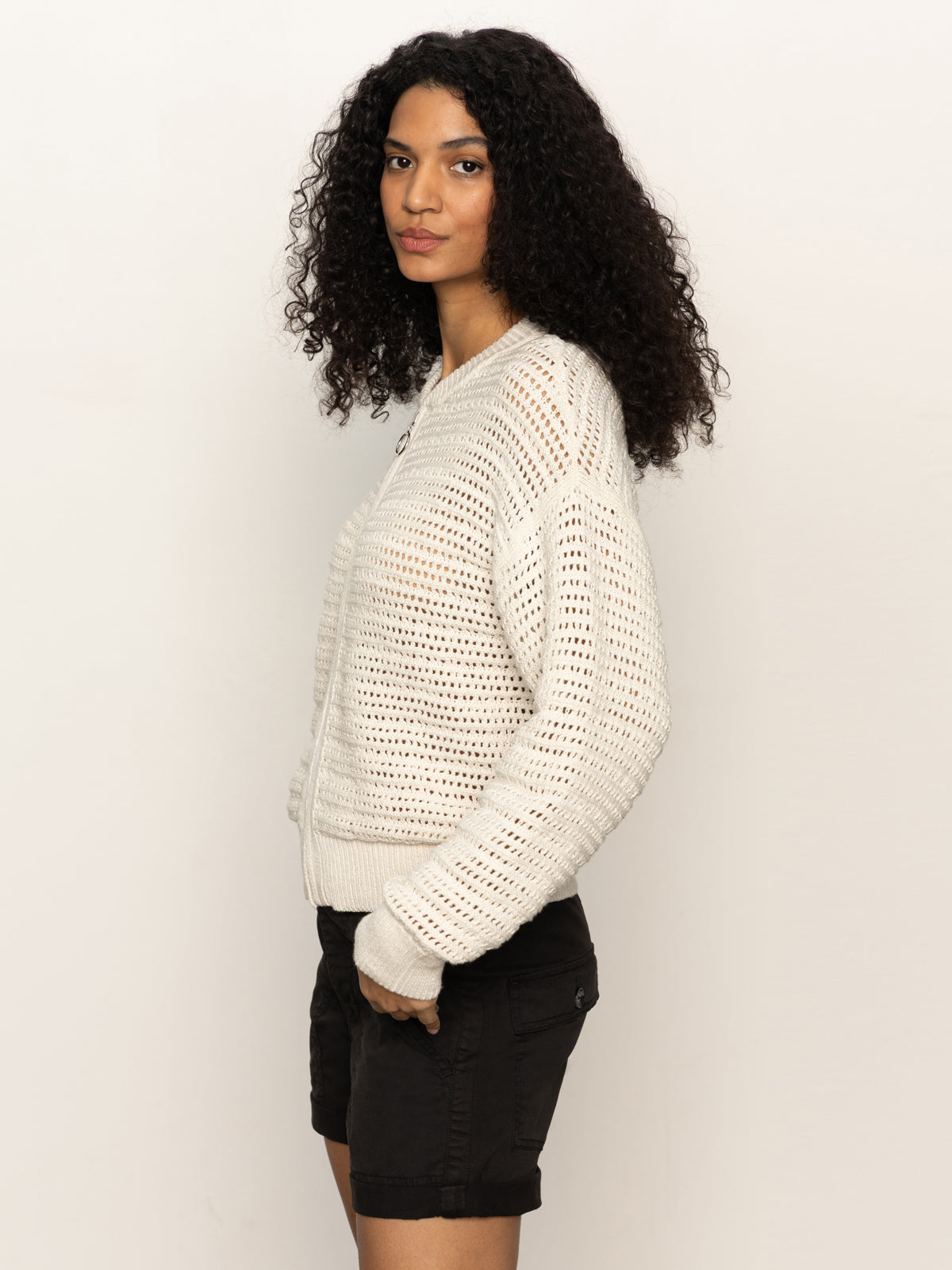 Woman with long curly hair wearing an open knit bomber jacket in chalk and black shorts from Sanctuary Clothing, standing sideways against a plain background.