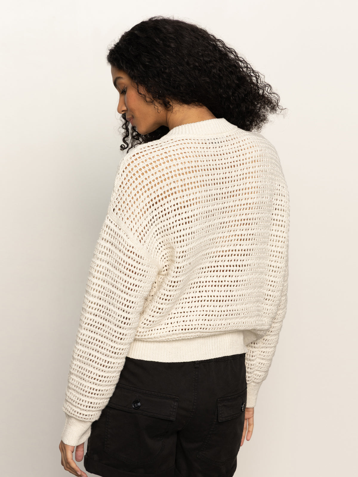 A person with curly hair is turned slightly away, showcasing a Sanctuary Clothing open knit bomber jacket in chalk. They are wearing black shorts, and the off-white background creates a soft contrast with the jacket's mesh-like pattern.