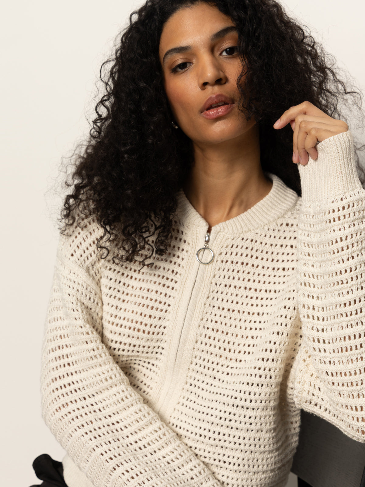 A person with long curly hair is wearing the open knit bomber jacket in chalk by Sanctuary Clothing while sitting on a chair against a plain white background, which highlights their relaxed pose and thoughtful expression.