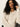 A person with long curly hair is wearing the open knit bomber jacket in chalk by Sanctuary Clothing while sitting on a chair against a plain white background, which highlights their relaxed pose and thoughtful expression.