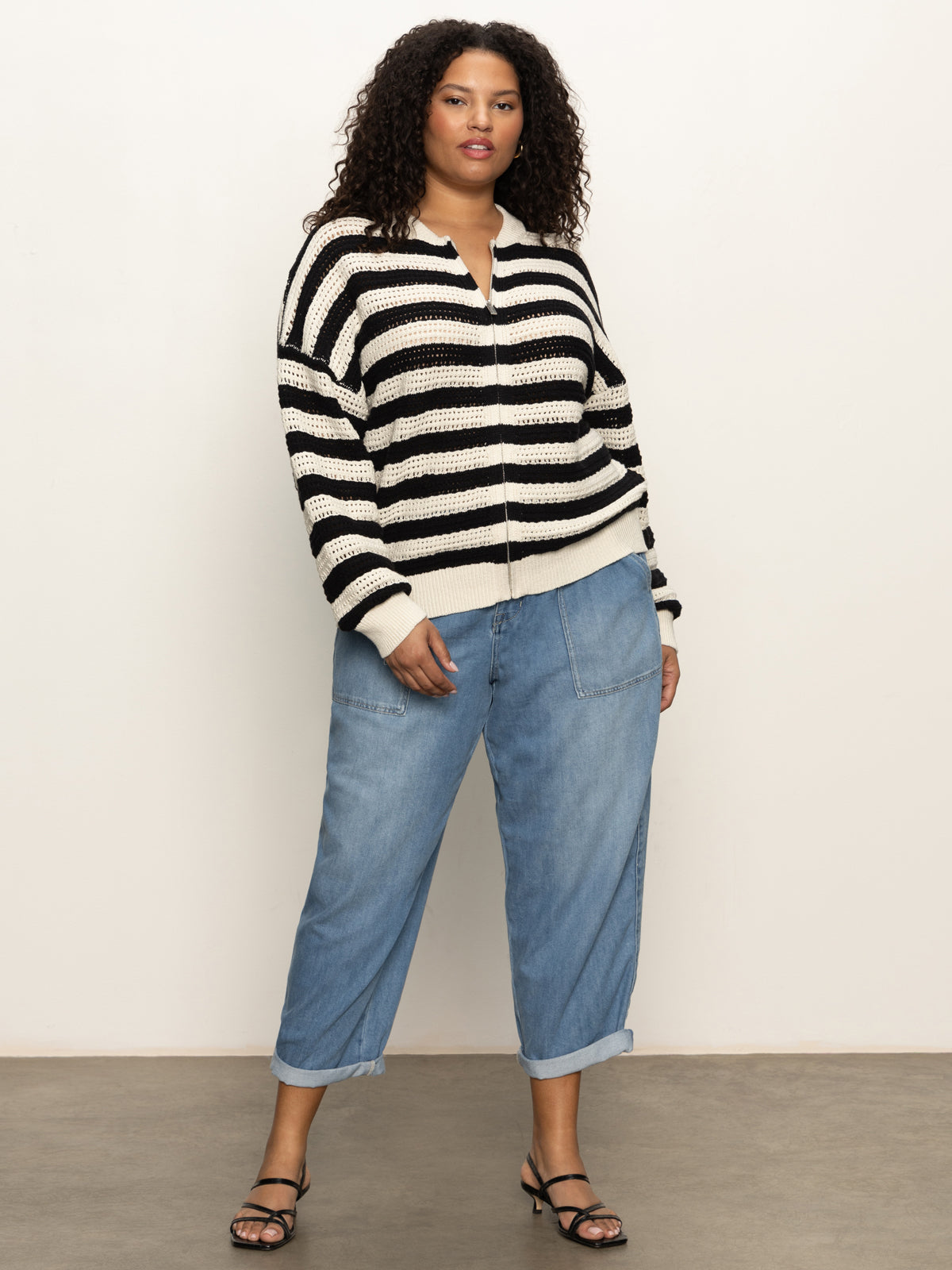 A woman with curly hair and a confident expression wears the Sanctuary Clothing open knit bomber jacket macrame black stripe (extended sizing) against a plain background, styled with light blue jeans and black sandals.