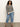 A woman with curly hair and a confident expression wears the Sanctuary Clothing open knit bomber jacket macrame black stripe (extended sizing) against a plain background, styled with light blue jeans and black sandals.