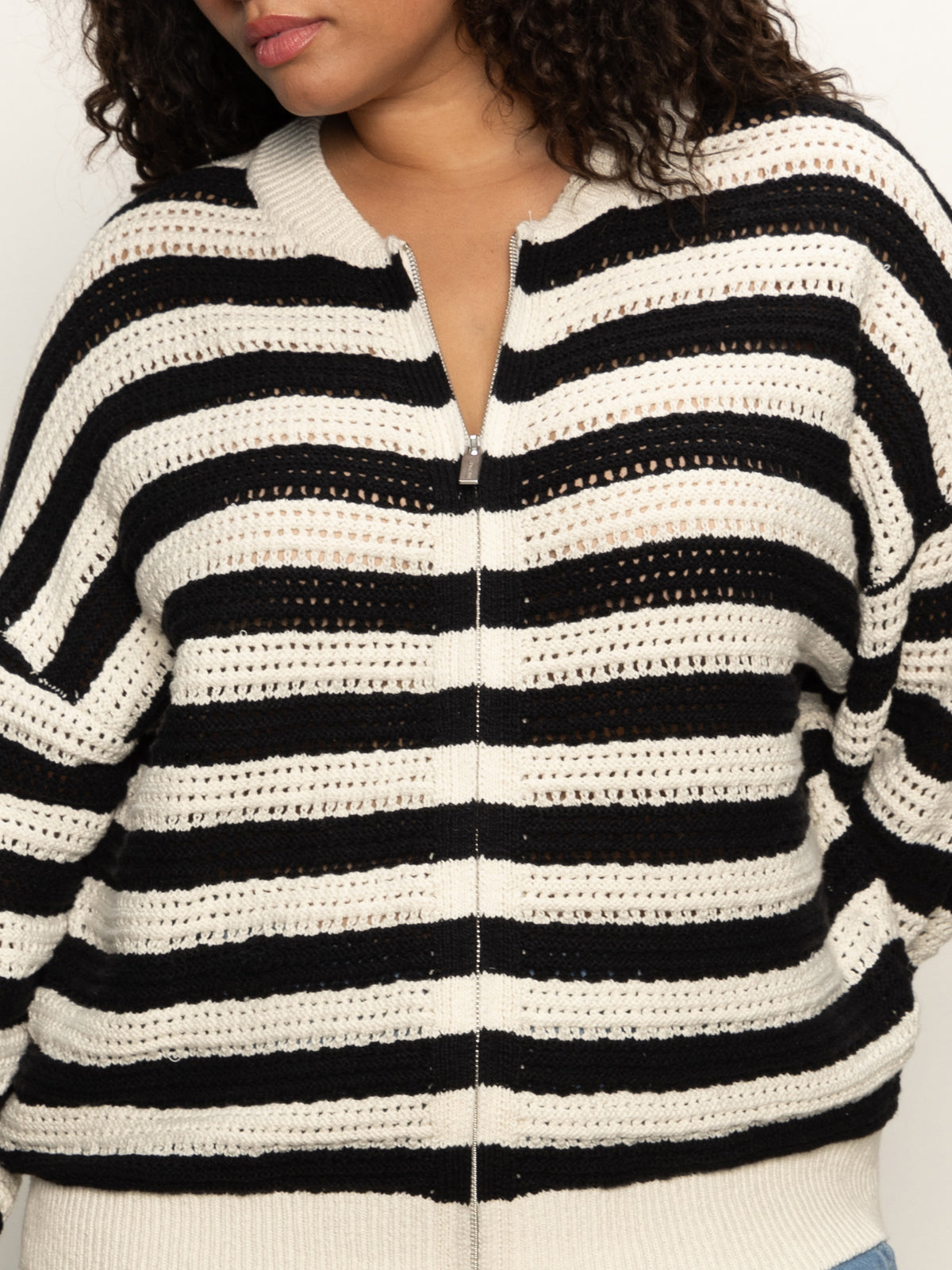 A person wears the Sanctuary Clothing open knit bomber jacket macrame black stripe (extended sizing), featuring black and white horizontal stripes, a zip front, loose fit, and textured open-knit pattern. Their face is partially out of frame.