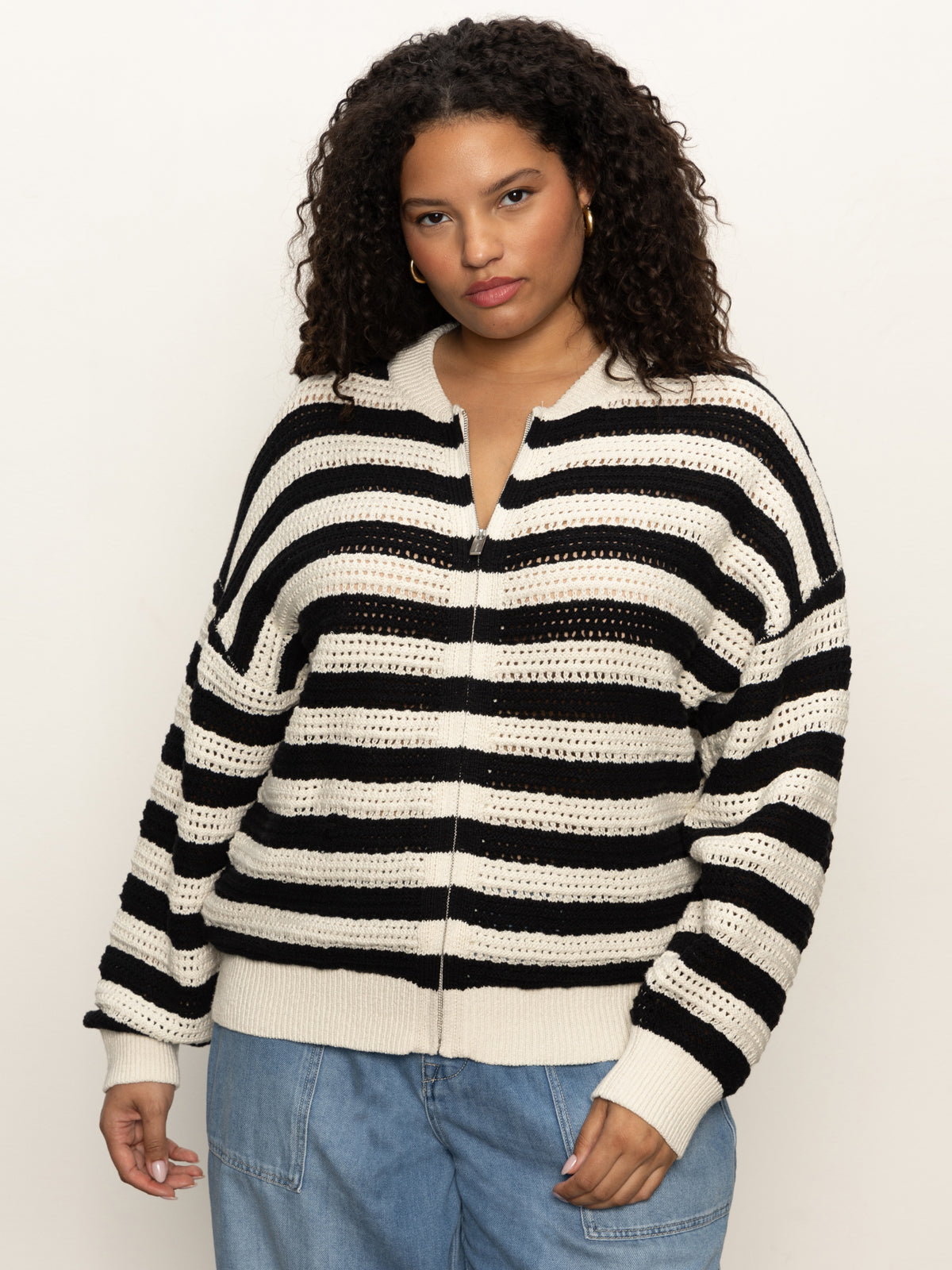 A woman with curly hair wears the Sanctuary Clothing open knit bomber jacket in black stripes (extended sizing), paired with light blue jeans, standing against a plain light background.