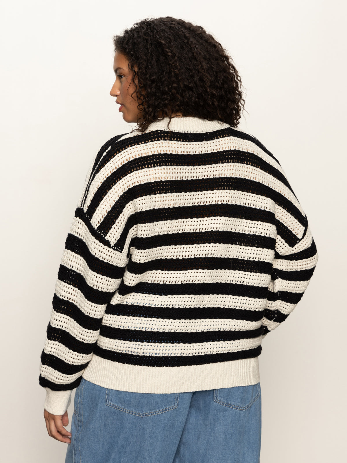 A person with curly hair is shown from the back, wearing the Sanctuary Clothing open knit bomber jacket macrame black stripe (extended sizing) and light blue denim pants against a plain light background.