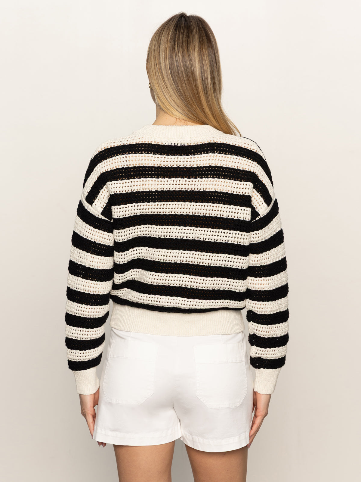 A woman with straight blonde hair wears the Sanctuary Clothing open knit bomber jacket macrame in black stripe with white shorts, facing away against a plain light background.