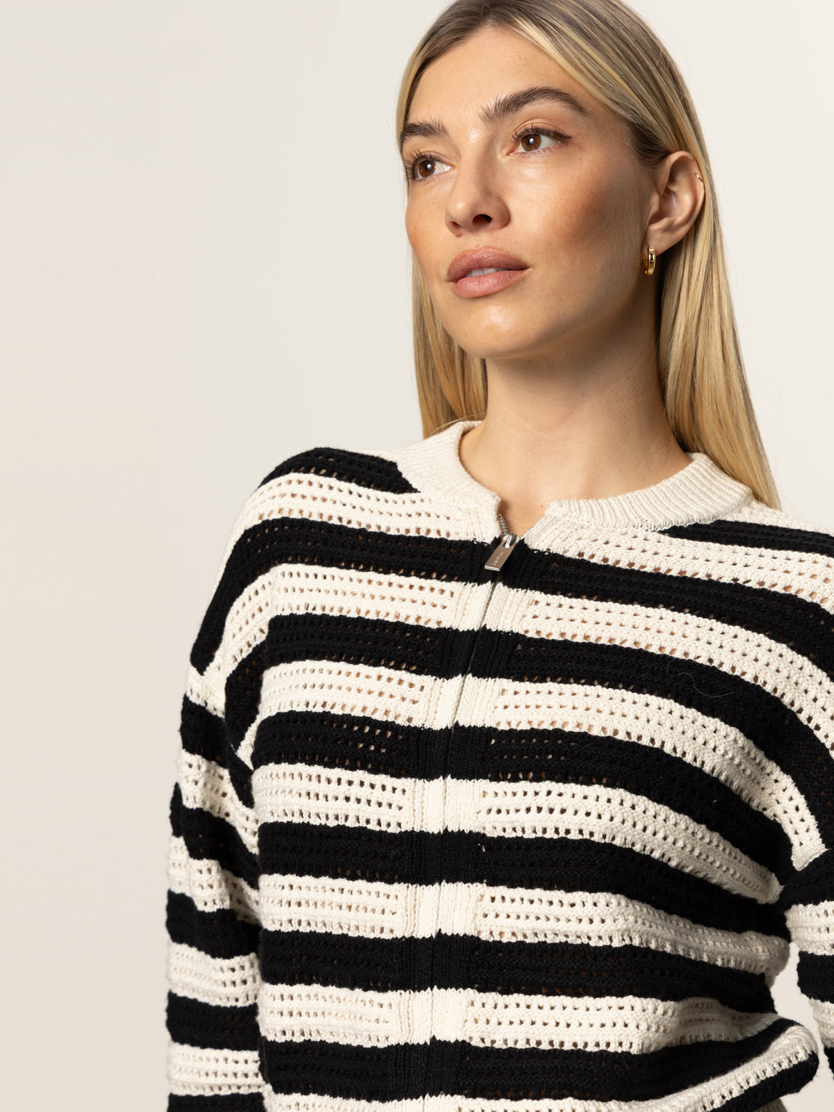 A woman with straight blonde hair models the Sanctuary Clothing open knit bomber jacket macrame black stripe, paired with gold hoop earrings, looking slightly upward against a plain light background.