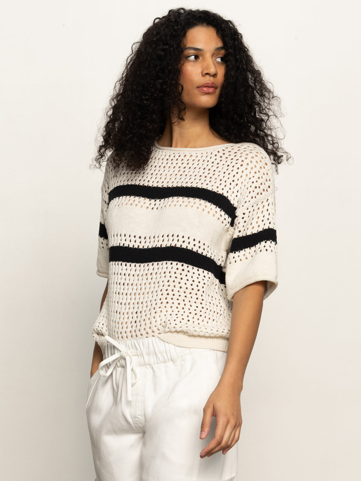 A woman with curly hair wears Sanctuary Clothing's "openwork tee black chalk stripe" over a white top, paired with light-colored pants. She stands against a plain background, looking to her left with one hand in her pocket.