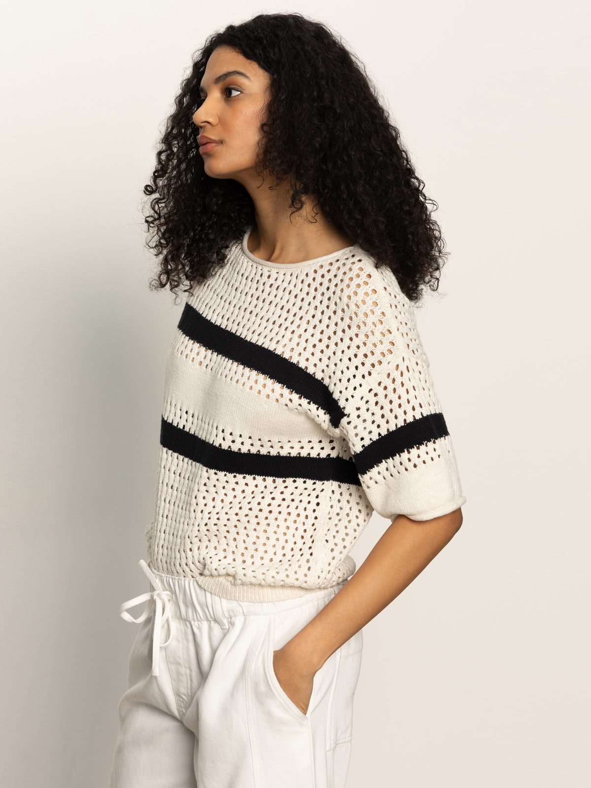 A person with curly hair is wearing Sanctuary Clothing's openwork tee in black chalk stripe, paired with white pants, standing sideways against a plain light background.
