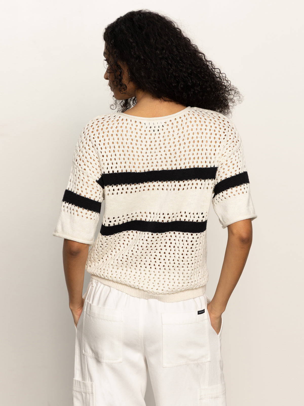 A person with curly hair, facing away, is wearing Sanctuary Clothing's openwork tee black chalk stripe with short sleeves and white pants. The background is plain white.